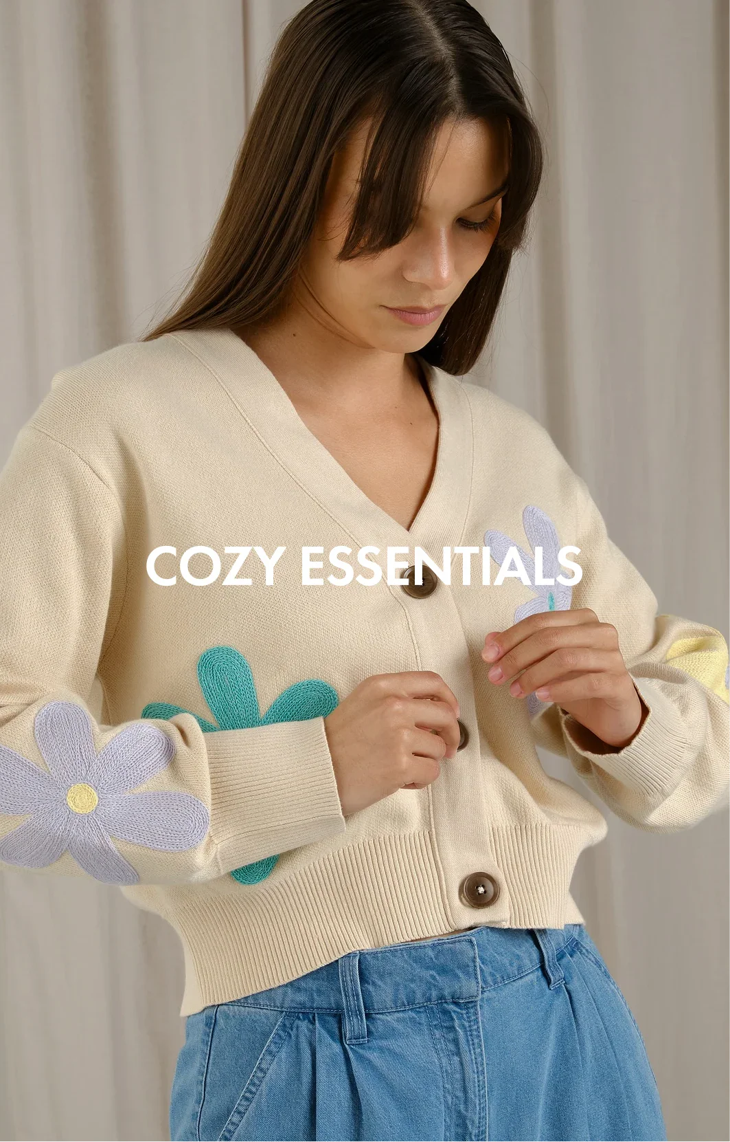 Woman wearing cream knit cardigan with large pastel flower patches and blue jeans, cozy essentials style.